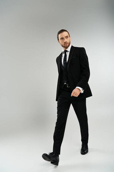 A bearded businessman confidently walks in a suit and tie against a grey studio backdrop.