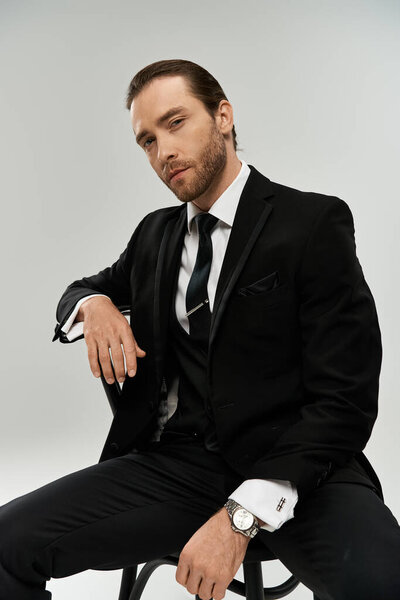 A handsome, bearded businessman sits on a chair in a studio, looking contemplative in his sleek suit against a grey background.