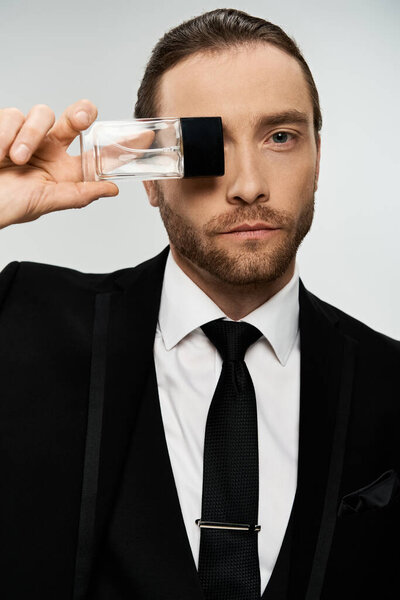 Handsome bearded businessman in a suit and tie carefully holding a glass perfume bottle on a grey studio background.