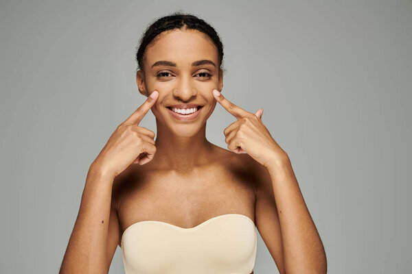 Young African American woman in a strapless bra smiling and pointing fingers to the side on a grey background.