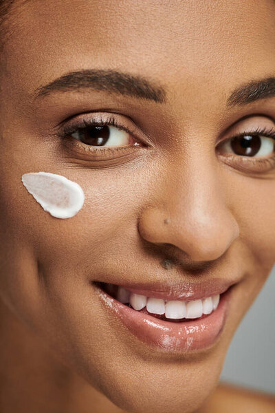Young African American woman in strapless top applying cream to her face, focusing on self-care, skincare routine.