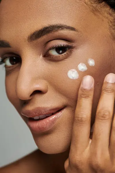 stock image Beautiful young African American woman in strapless top applying a thick layer of cream to her face on a grey background.