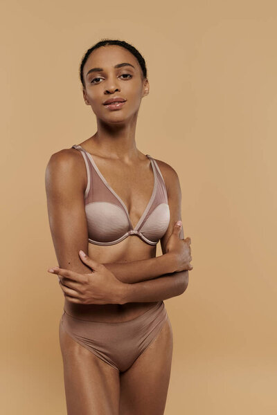 A pretty, slim African American woman poses confidently in tan lingerie on a beige background.