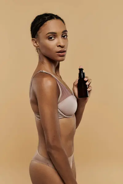 stock image A slim African American woman in a bikini enjoys a bottle of body oil against a beige backdrop.