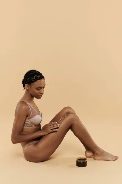 Slim African American woman in underwear sits gracefully with coffee scrub on legs on a beige background.