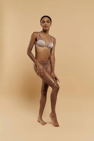 Beautiful, slim African American woman in a bikini confidently posing with coffee scrub on legs on a beige background.