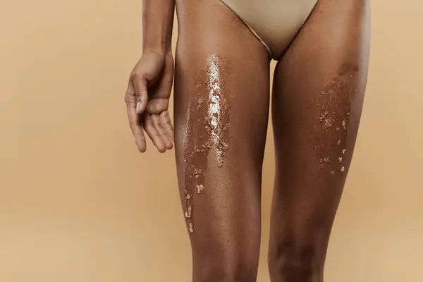 Stock image African American woman showcasing her beauty in a bikini coated with sugar scrub on legs on a beige backdrop.