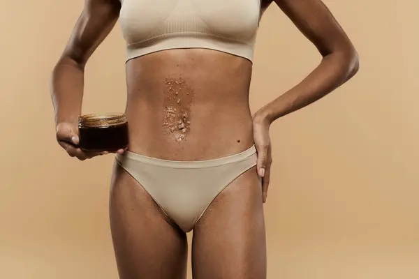 stock image A slender African American woman in a bikini holding a jar of scrub, caring for her body against a beige background.