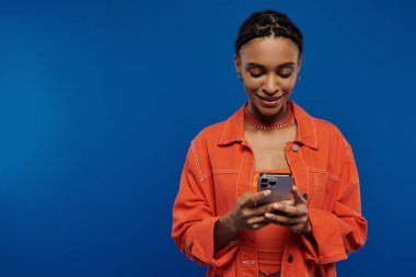 Turuncu gömlekli genç bir Afrikalı Amerikalı kadın cep telefonuna odaklanmış durumda..