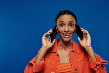 Canlı, turuncu elbiseli Afrikalı Amerikalı bir kadın iki cep telefonunu kulağına dayayıp teknolojiyle bağlantı kuruyor..