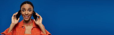 Young African American woman in vibrant orange outfit holding two cell phones to her ears on a blue background. clipart