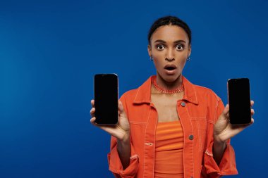 Canlı turuncu kıyafetli genç Afrikalı Amerikalı kadın mavi arka planda iki cep telefonu tutuyor..