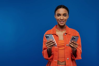 Turuncu elbiseli şık bir Afrikalı Amerikalı kadın mavi arka planda iki cep telefonu tutuyor..