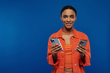 A stylish African American woman in orange holds two cell phones against a blue background. clipart