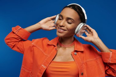 Turuncu tişörtlü genç Afrikalı Amerikalı kadın mavi arka planda kulaklıkla müzik dinliyor..