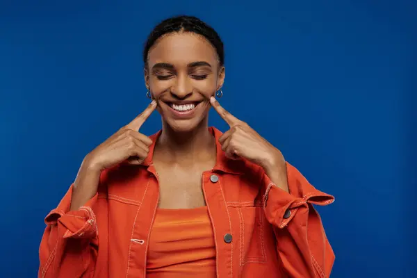 stock image A pretty young African American woman in a vibrant orange shirt on a blue background making a funny face.