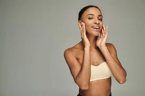 stock image Young African American woman smiling, hands to face, wearing a bra top, radiating joy and self-care on grey background.