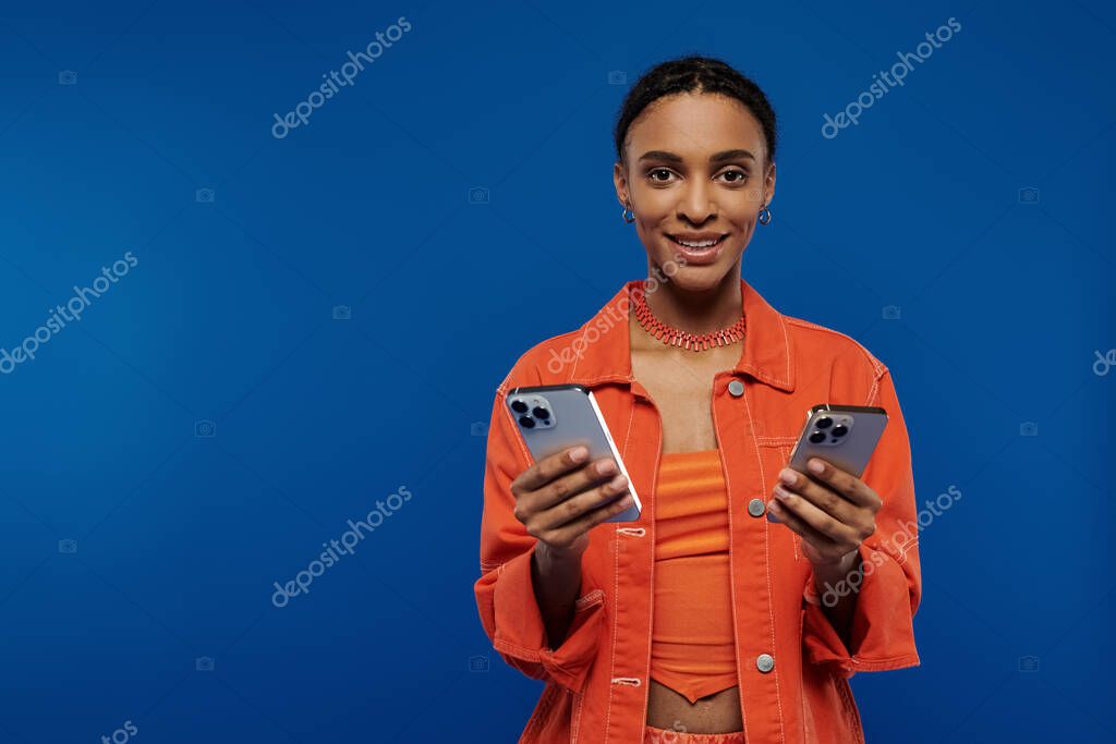 A stylish African American woman in orange holds two cell phones against a blue background.