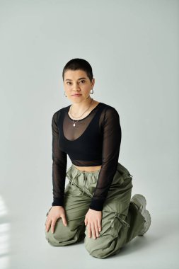 Young woman with short hair striking a pose in a black top and green pants, against a grey background. clipart