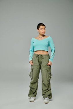 Kısa saçlı, modaya uygun genç bir kadın, mavi bluzlu ve kargo pantolonlu bir pozu parlak gri arka planda karşılıyor..