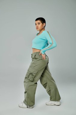 Kısa saçlı genç kadın, modaya uygun mavi bluzlu ve haki pantolonlu pozlar veriyor. Tarafsız gri bir zemine karşı..