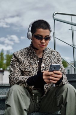 Leopar desenli ceketli şık bir kadın basamaklara oturmuş kulaklık takıyor ve cep telefonuna bakıyor..