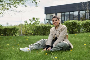 A young woman with short hair sits on lush green grass in front of a modern building. clipart