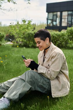 Kısa saçlı şık bir kadın çimlerin üzerinde oturuyor, akıllı telefon ekranına odaklanmış..