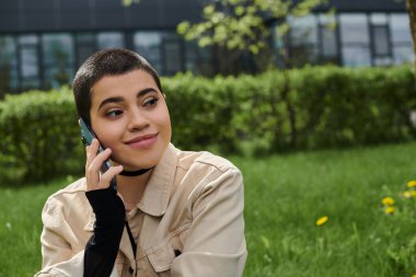 A young woman with short hair smiling as she talks on smartphone while sitting on green grass outdoors. clipart