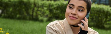 Bir tarlada oturan genç bir kadın, cep telefonuyla konuşuyor, doğanın güzelliği ve yeşil çimlerle çevrili..