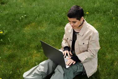 Kısa saçlı genç bir kadın çimlerin üzerinde şık bir şekilde rahat bir şekilde oturur ve açık havada dizüstü bilgisayar kullanmaya odaklanır..