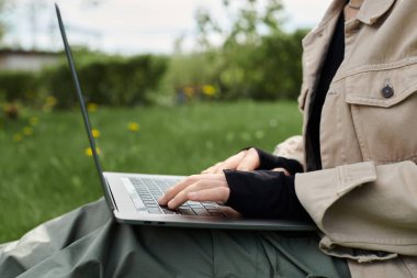 Genç bir kadın yeşil çimenlerde dizüstü bilgisayar kullanarak şık bir şekilde oturuyor..