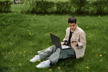 Çimenlerde oturan bir çocuk, gözleri bilgisayar ekranına odaklanmış, dijital dünyayı keşfederken doğa tarafından çevrelenmiş..