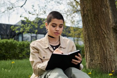 A young woman with short hair immersed in a book while seated stylishly on green grass outdoors. clipart