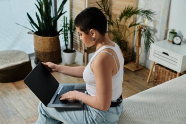 A young woman with short hair in casual attire sitting on a bed, engrossed in using a laptop computer. clipart