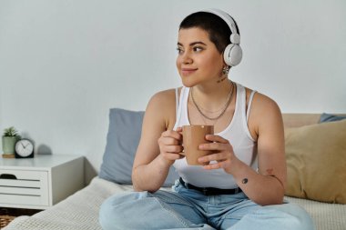 Casual young woman with short hair sitting on bed, enjoying a cup of coffee. clipart