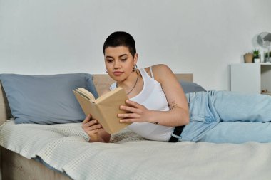 Kısa saçlı, gündelik giyinmiş genç bir kadın, bir yatağa uzanıyor, kitap okumaya dalmış..