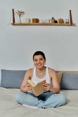 A young woman with short hair sits on a bed engrossed in a book, peaceful and absorbed in her reading. clipart