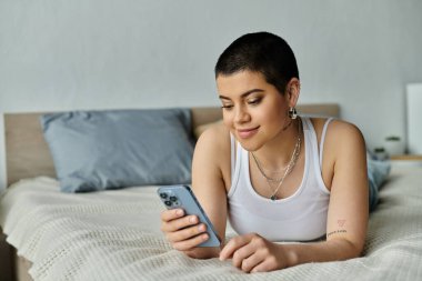 Kısa saçlı genç bir kadın cep telefonu ekranına dalmış bir yatakta dinleniyor..