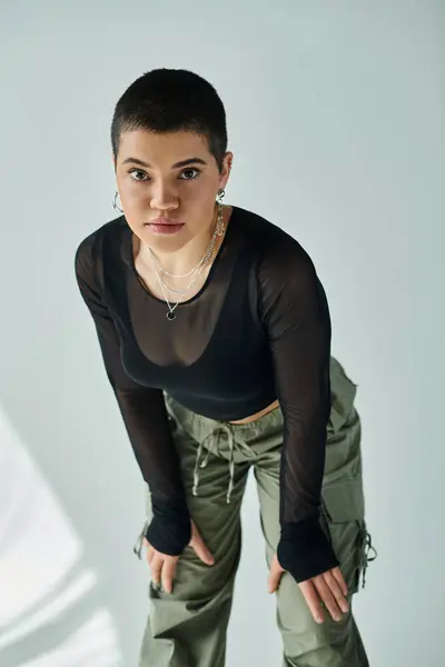 stock image Young woman with short hair strikes a pose in fashionable black shirt and green pants on a grey background.