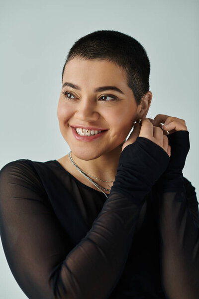 Молодая женщина с короткими волосами ярко улыбается, излучая уверенность в стильном черном платье на сером фоне.