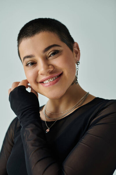 Молодая женщина с короткими волосами, одетая в шикарный черный топ, ярко улыбающаяся, позируя на сером фоне.