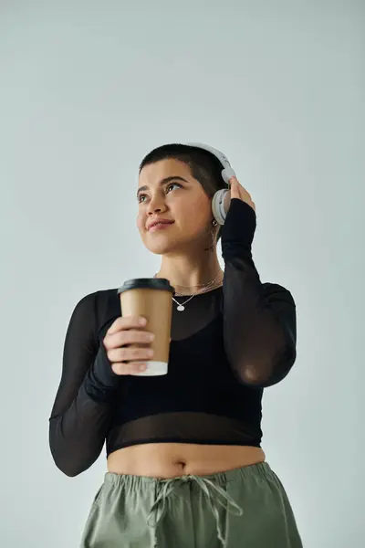 stock image A young woman with short hair in casual attire multitasking with a cup of coffee and a cell phone on a grey background.