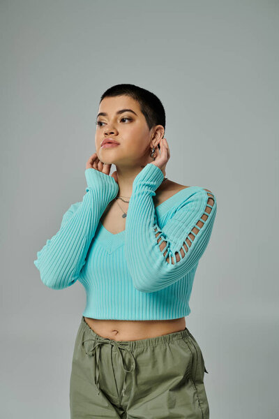 A young woman with short hair striking a pose in a stylish blue sweater and green pants against a grey backdrop.