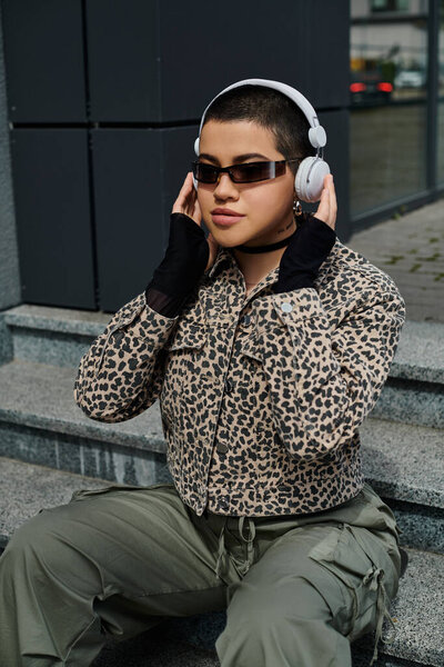 Стильная молодая женщина с короткими волосами сидит на ступеньках в наушниках перед современным зданием, под музыку.