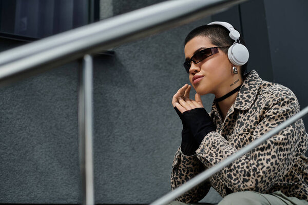 Стильно одетая молодая женщина с короткими волосами в наушниках, сидящая на городских ступеньках возле современного здания.