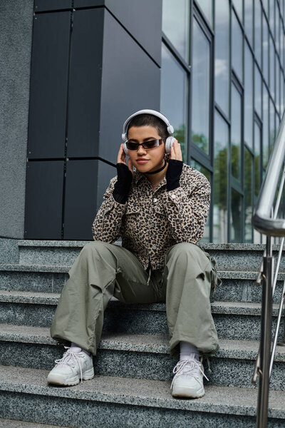 Мужчина в наушниках сидит на ступеньках, затерянный в музыке, рядом со стильной женщиной в леопардовой куртке у современного здания.