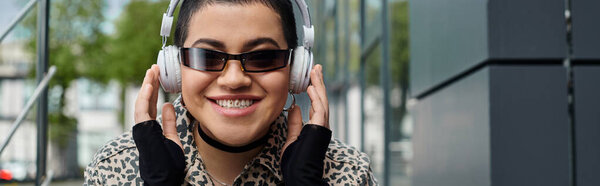 Стильная молодая женщина с короткими волосами в наушниках и леопардовую рубашку рядом с современным зданием.