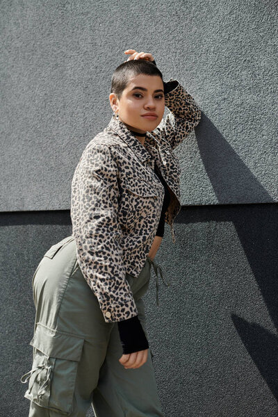 A stylish young woman in a leopard print jacket leans against a modern building, hand on head.