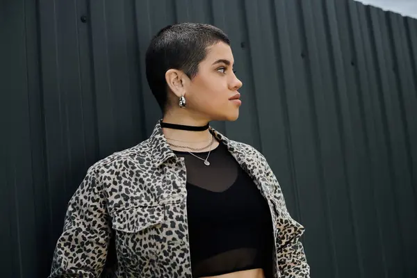 stock image A stylish young woman with short hair wears a black top and a leopard print jacket near a modern building.